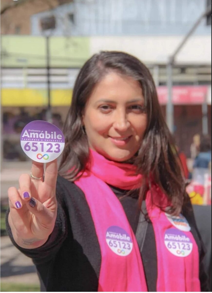 Amábile Marchi votando em 2024, de camiseta branca com símbolo do infinito da neurodiversidade, fazendo sinal de vitória